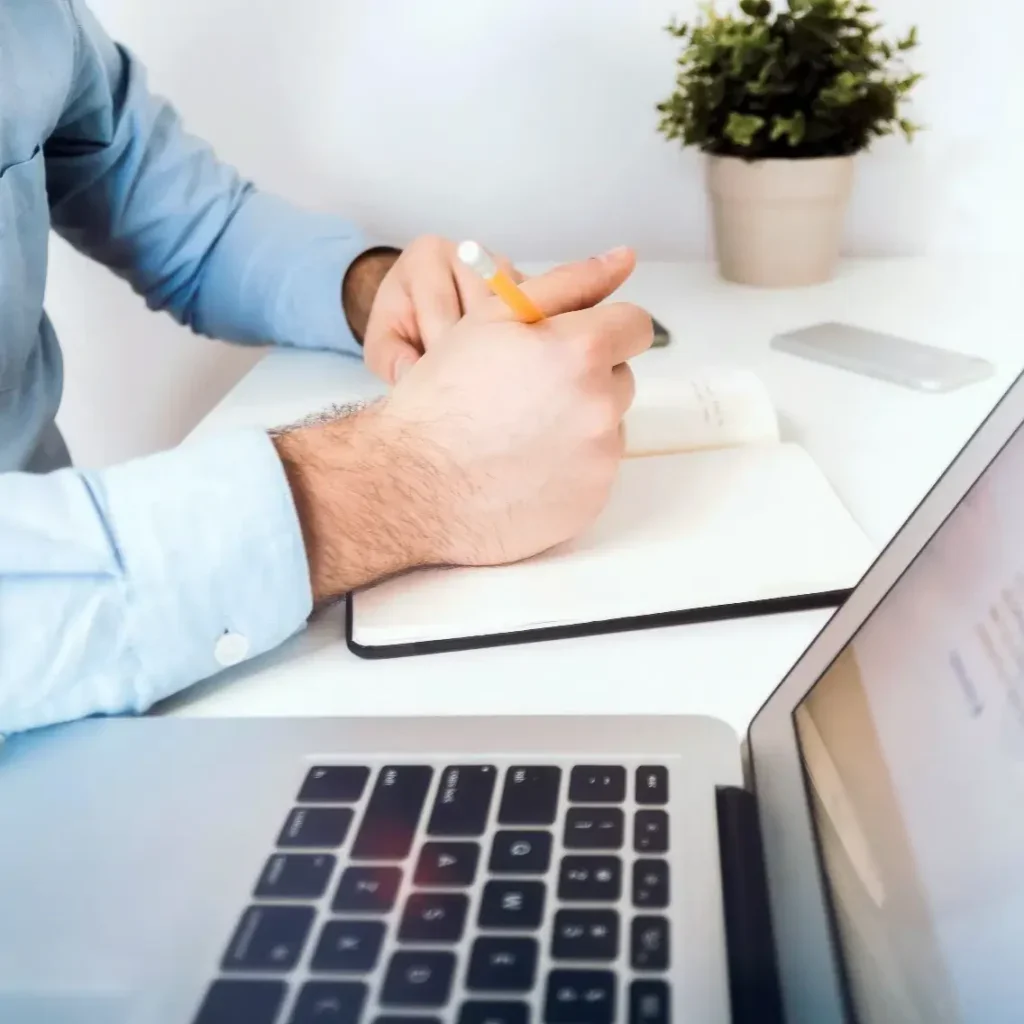 Man taking notes in a journal next to a laptop computer.